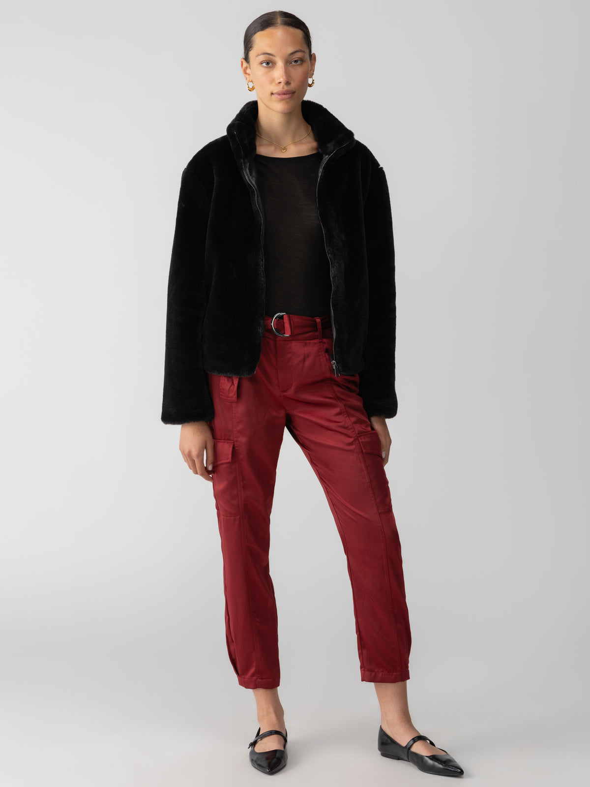 A person is posing against a plain background, wearing the Lux Fur Coat Black by Sanctuary Clothing over a black top, paired with red cargo pants and black flats. They have their hair pulled back and are accessorized with gold hoop earrings. Their expression is neutral.