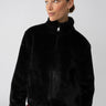 A person wearing the Lux Fur Coat Black by Sanctuary Clothing and red pants poses against a plain background, gazing to the right. The coat features a high collar, and the person has neatly pulled-back hair and is wearing hoop earrings.