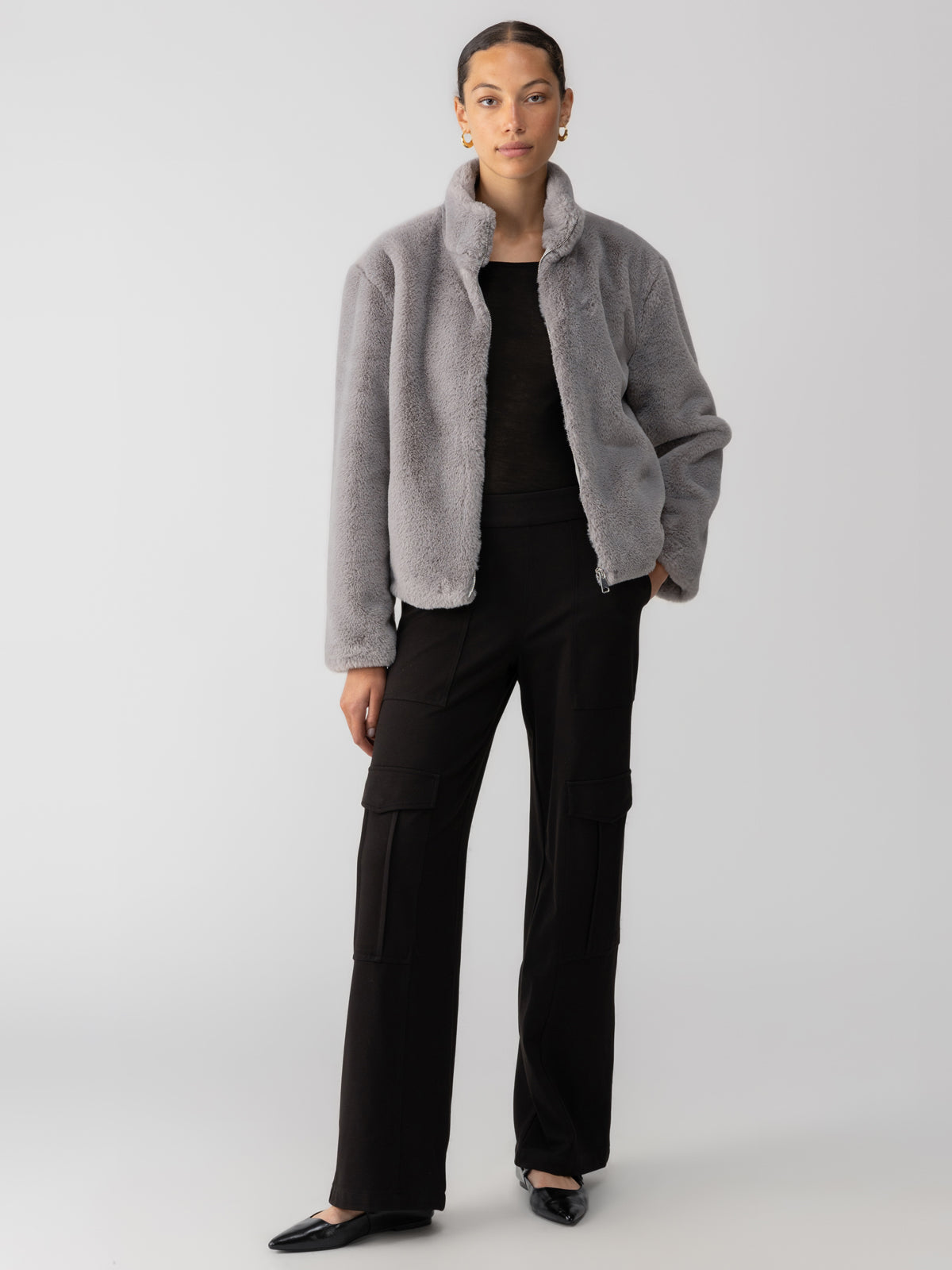 A person stands against a plain background wearing the Lux Fur Coat Silver Fox from Sanctuary Clothing over a black top and black cargo pants. They have earrings and black shoes. The person has their hair pulled back and one hand in their coat pocket, striking a confident pose.