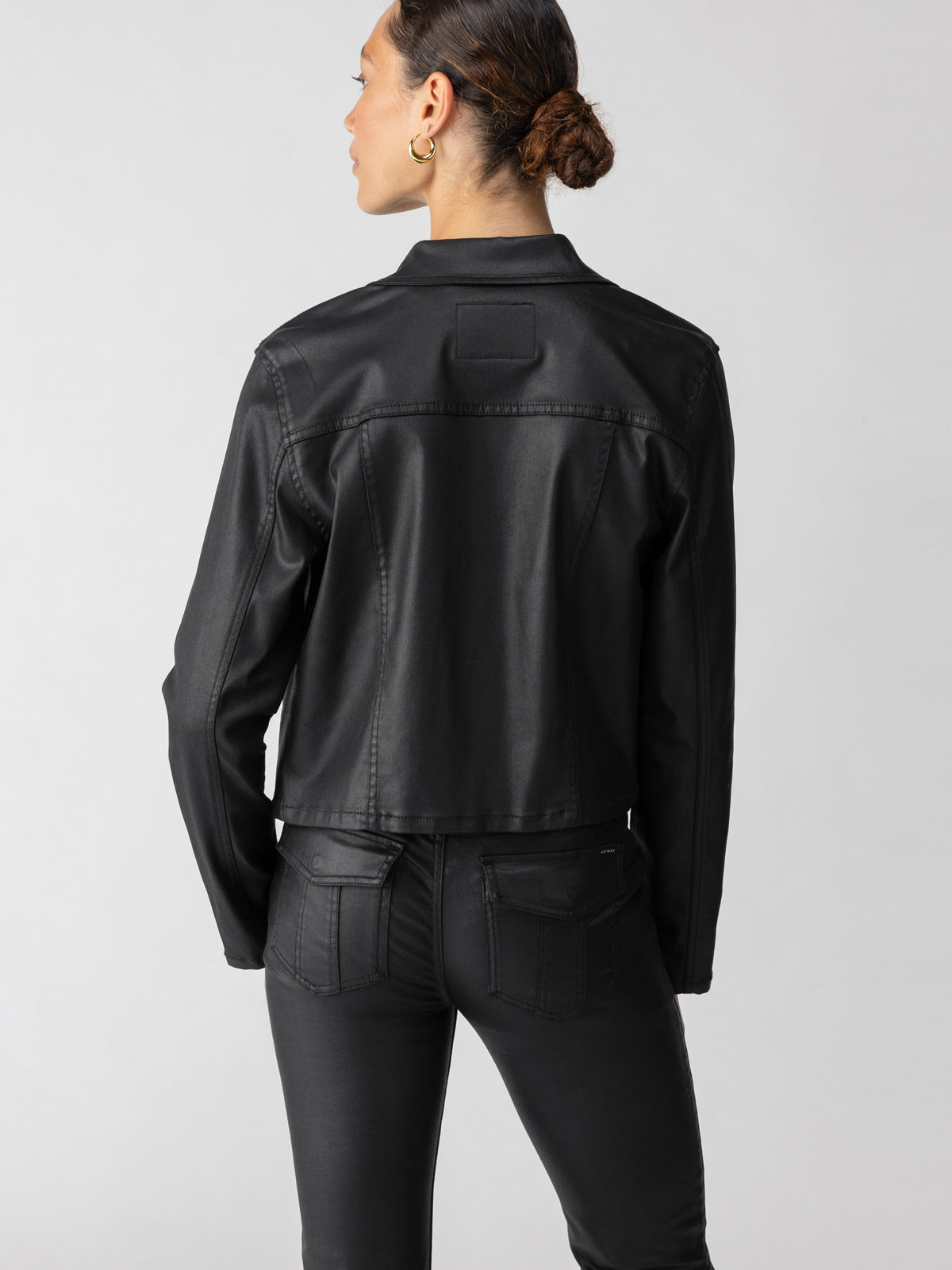 A person with a neat bun hairstyle is standing with their back to the camera, showcasing the stylish Davidson Trucker Jacket Black by Sanctuary Clothing and matching black pants. The plain, light-colored background draws attention to the clothing.