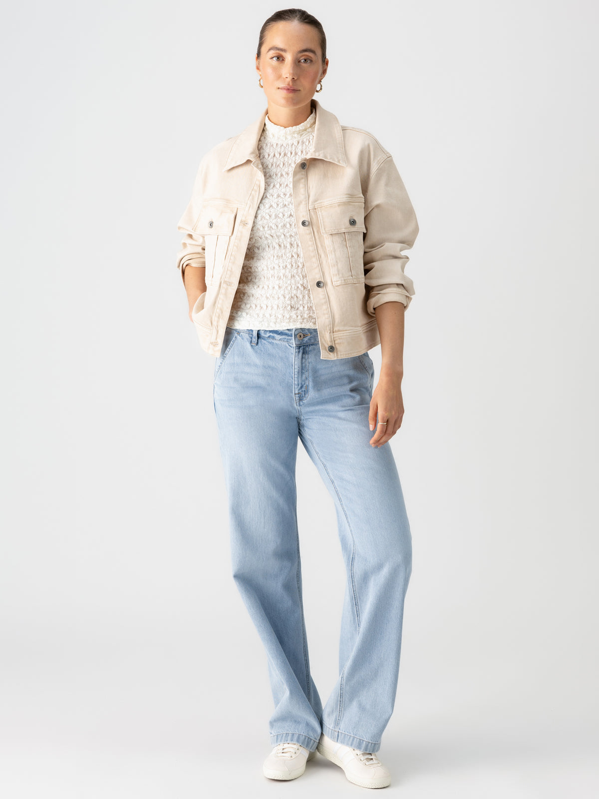 A person stands confidently against a plain background wearing the Cargo Denim Jacket in Toasted Almond from Sanctuary Clothing over a white knit sweater, light blue high-waisted jeans, and white sneakers. They have their hands in their jacket pockets and a calm expression on their face.