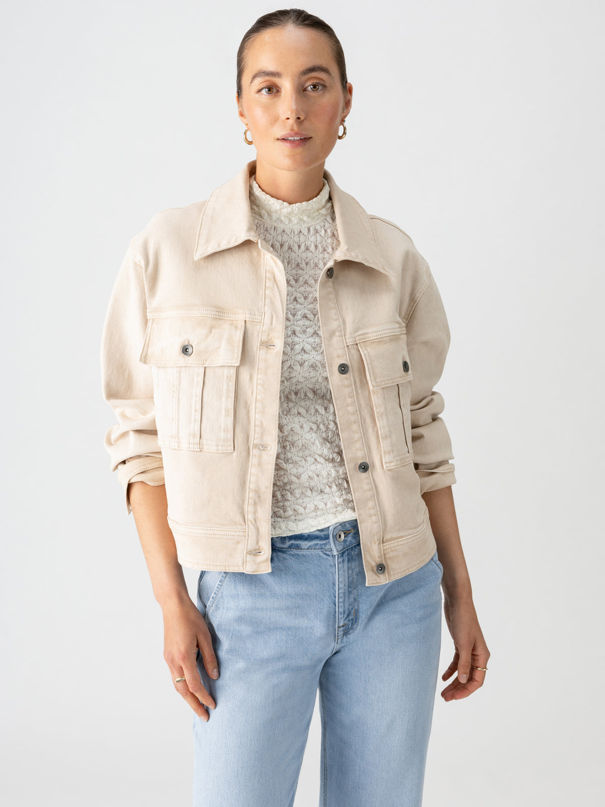 A person wearing the Cargo Denim Jacket Toasted Almond by Sanctuary Clothing over a white textured sweater and light blue jeans stands against a plain background. The person has their hair pulled back and is looking directly at the camera, with one hand by their side and the other lightly touching their jacket.