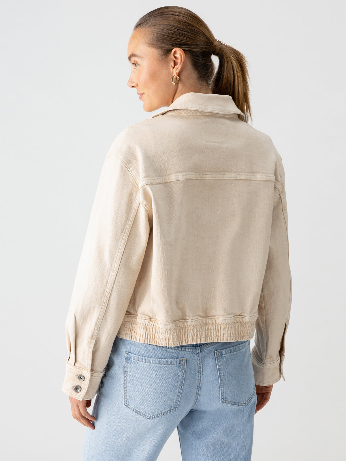 A woman with light brown hair tied in a ponytail is shown from the back, wearing a Sanctuary Clothing Cargo Denim Jacket in Toasted Almond and blue jeans. She has gold hoop earrings and stands against a plain, light background.