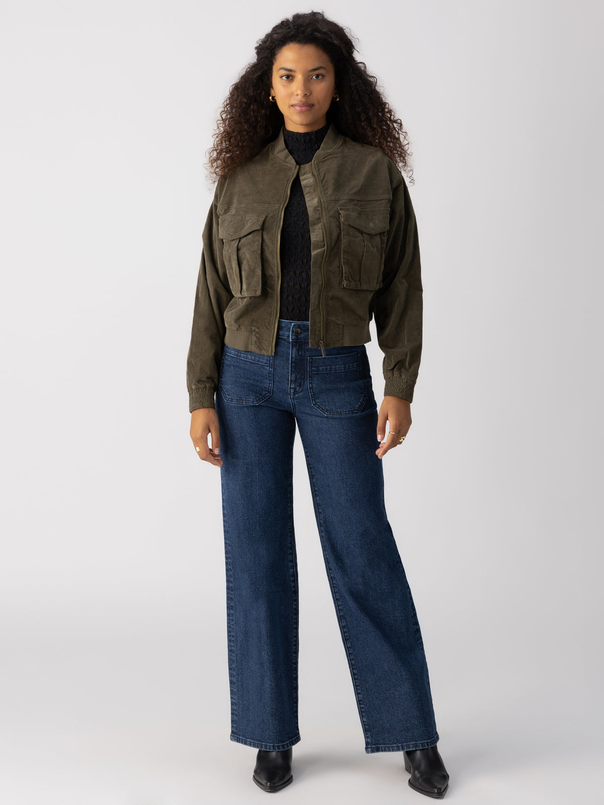 A woman with long, curly hair is wearing the Corduroy Surplus Bomber in Burnt Olive by Sanctuary Clothing over a black top, paired with high-waisted blue wide-leg jeans and black boots. She stands against a plain, light gray background and looks towards the camera with a calm expression.