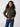 A woman with long curly hair wears the Corduroy Surplus Bomber in Burnt Olive by Sanctuary Clothing paired with blue jeans. She stands against a plain, light-colored background, looking confidently at the camera with her hands in her jacket pockets.