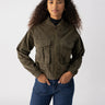 A woman with long curly hair wears the Corduroy Surplus Bomber in Burnt Olive by Sanctuary Clothing paired with blue jeans. She stands against a plain, light-colored background, looking confidently at the camera with her hands in her jacket pockets.