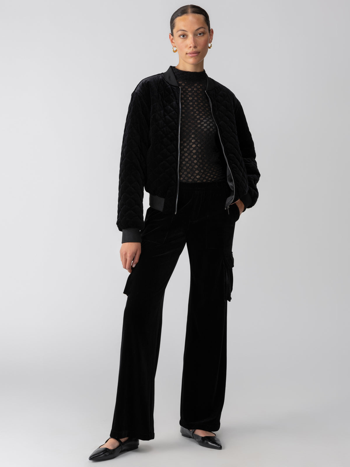 A person stands confidently against a plain background. They are wearing the Mona Velvet Bomber Black by Sanctuary Clothing over a sheer black top, paired with black wide-leg trousers. They have their hands in their pockets and are wearing black shoes.