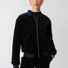 A person stands against a plain background wearing the Mona Velvet Bomber Black by Sanctuary Clothing. The jacket features a zip-up front and ribbed cuffs and collar. They have short hair pulled back, are wearing gold hoop earrings, and have a confident expression. Their hands are relaxed in the jacket pockets.