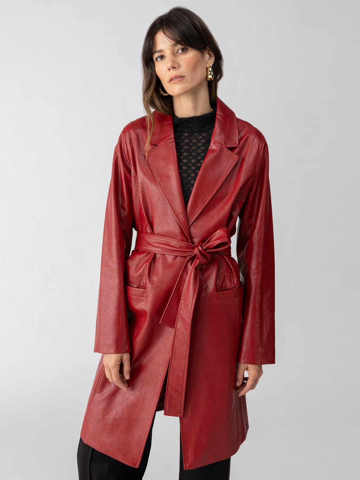 A woman with long dark hair is wearing the Vegan Leather Coat Garnet by Sanctuary Clothing, belted at the waist over a black lacy top. She stands against a plain white background and looks slightly to the side with a neutral expression.