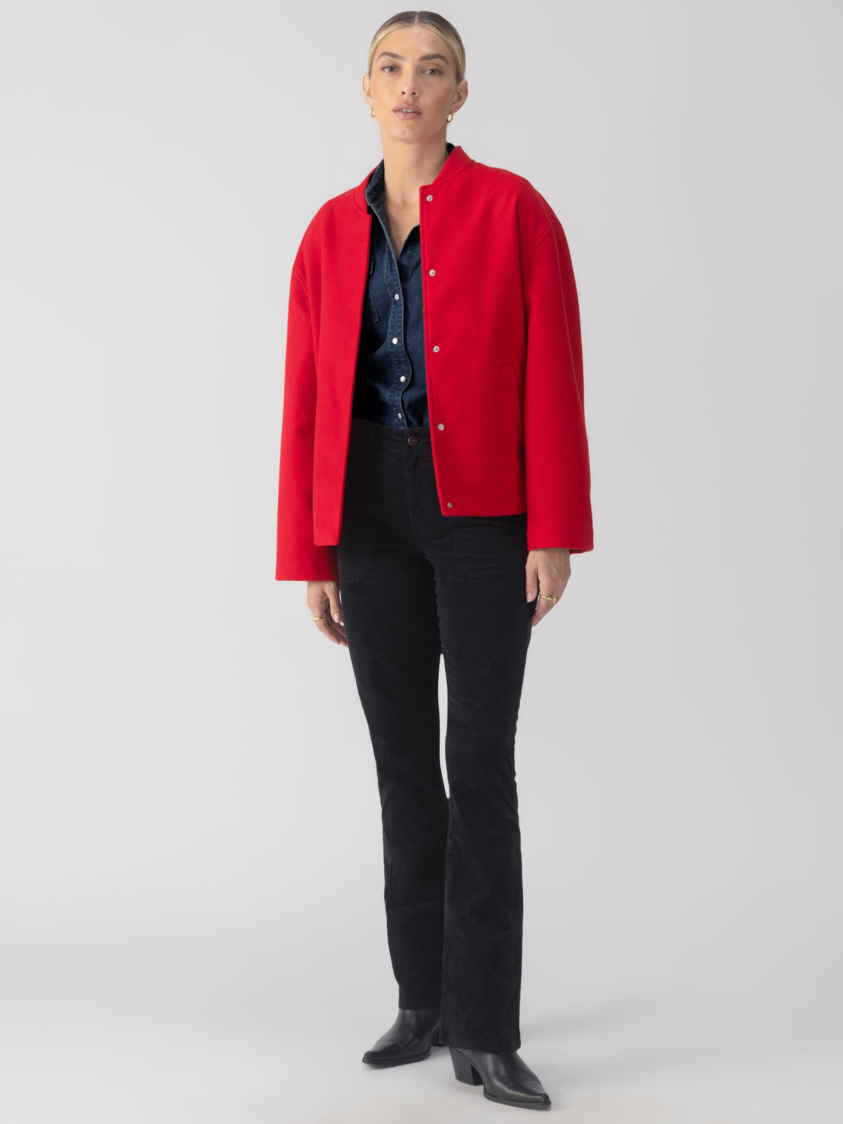 A person stands against a plain background wearing the Modern Bomber Jacket Mars Red from Sanctuary Clothing over a dark blue denim shirt and black trousers. They have on black ankle boots, and their hair is styled back.