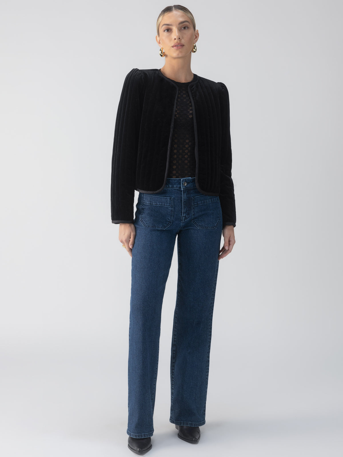 A person stands against a plain background wearing the Quilted Velvet Jacket in black from Sanctuary Clothing, a black top with a subtle pattern, blue jeans, and black shoes. They have short hair and are wearing earrings.