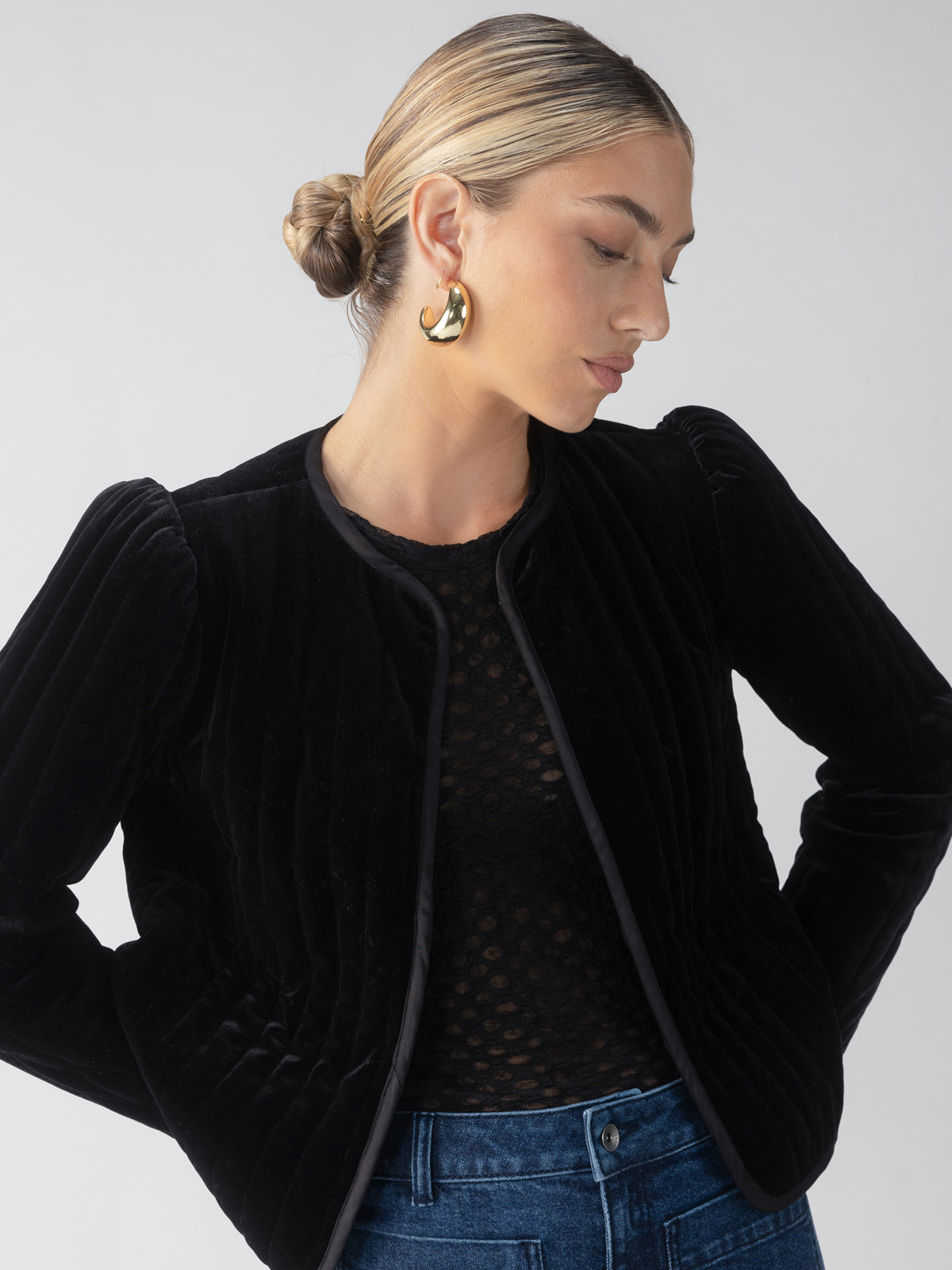 A woman with a sleek bun is wearing the Sanctuary Clothing Quilt Velvet Jacket in black over a sheer black top and blue jeans. She accessorizes with large gold hoop earrings, posing against a neutral background.