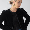 A woman with a sleek bun is wearing the Sanctuary Clothing Quilt Velvet Jacket in black over a sheer black top and blue jeans. She accessorizes with large gold hoop earrings, posing against a neutral background.