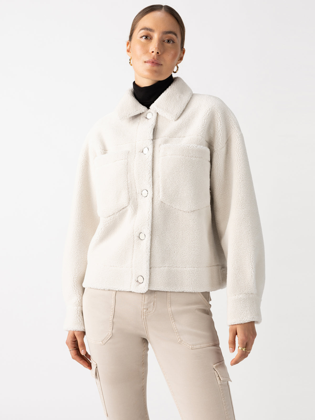 The individual is wearing the Cozy Sherpa Shacket Chalk from Sanctuary Clothing, featuring a light-colored, textured design with two prominent front pockets. Paired with beige pants, the shacket is buttoned up as the person poses against a plain backdrop, hair pulled back neatly.