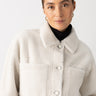 A person is wearing the "Cozy Sherpa Shacket Chalk" by Sanctuary Clothing, layered over a black turtleneck. Their hair is neatly tied back, and they are accessorized with gold hoop earrings against a plain white background.