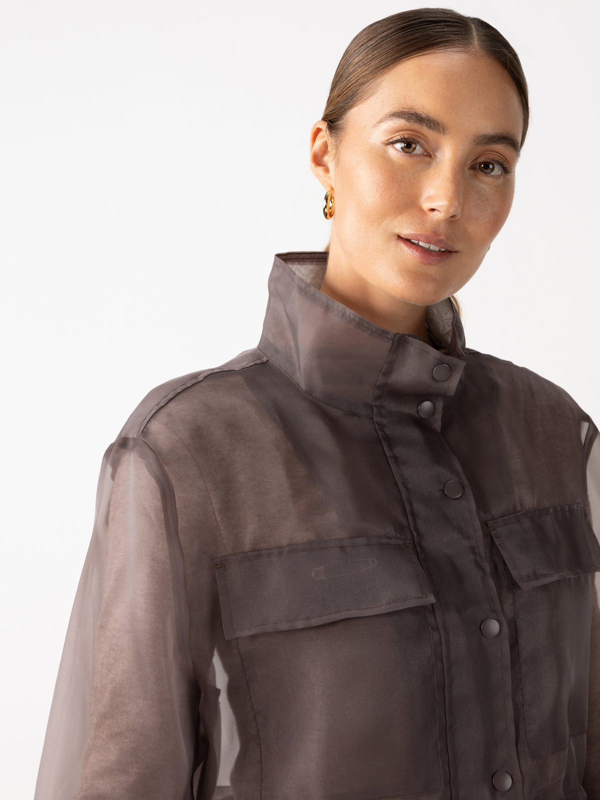 A person is wearing an organza parka in a cocoa hue from Sanctuary Clothing, featuring a transparent design, large pockets, and a button-up front. With their hair pulled back and wearing small hoop earrings, the person smiles slightly against a plain white background.