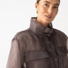 A person is wearing an organza parka in a cocoa hue from Sanctuary Clothing, featuring a transparent design, large pockets, and a button-up front. With their hair pulled back and wearing small hoop earrings, the person smiles slightly against a plain white background.