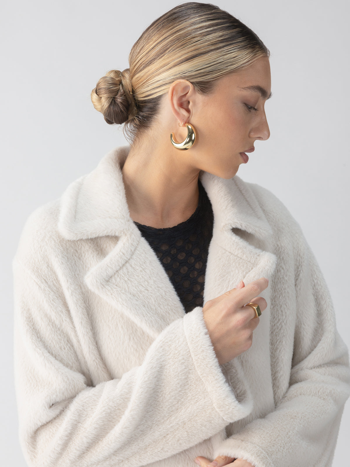 A woman with a sleek bun hairstyle gazes to the side, wearing Sanctuary Clothing's Live In Fur Coat Frosted White over a black top with a subtle pattern. Large gold hoop earrings and a matching ring complement her look against a neutral gray background.