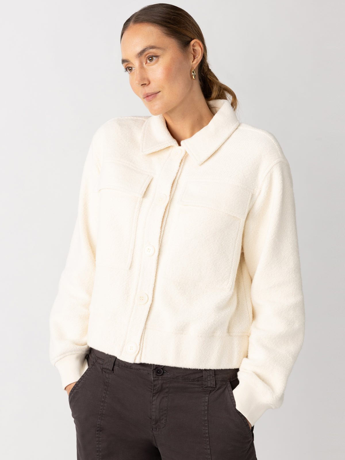 A person wearing the "cruise knit jacket chalk" by Sanctuary Clothing, which features a cream color and a button-up design with a collar, stands against a plain background. They have light brown hair pulled back and are wearing dark pants, while looking slightly to the side with a neutral expression.