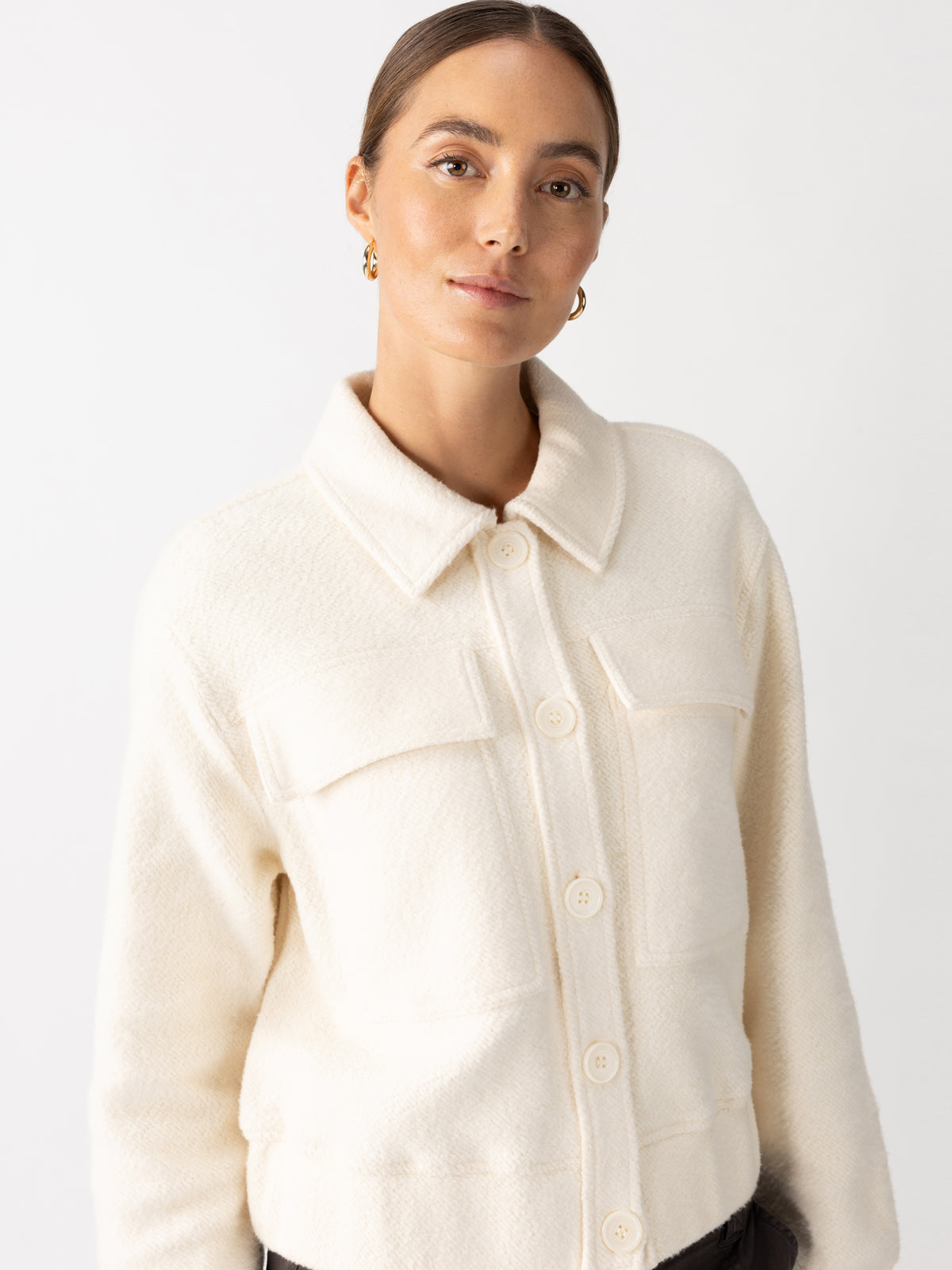 Wearing the Cruise Knit Jacket Chalk by Sanctuary Clothing, a person gazes softly at the camera. Their hair is pulled back and they sport small hoop earrings against a plain white background.