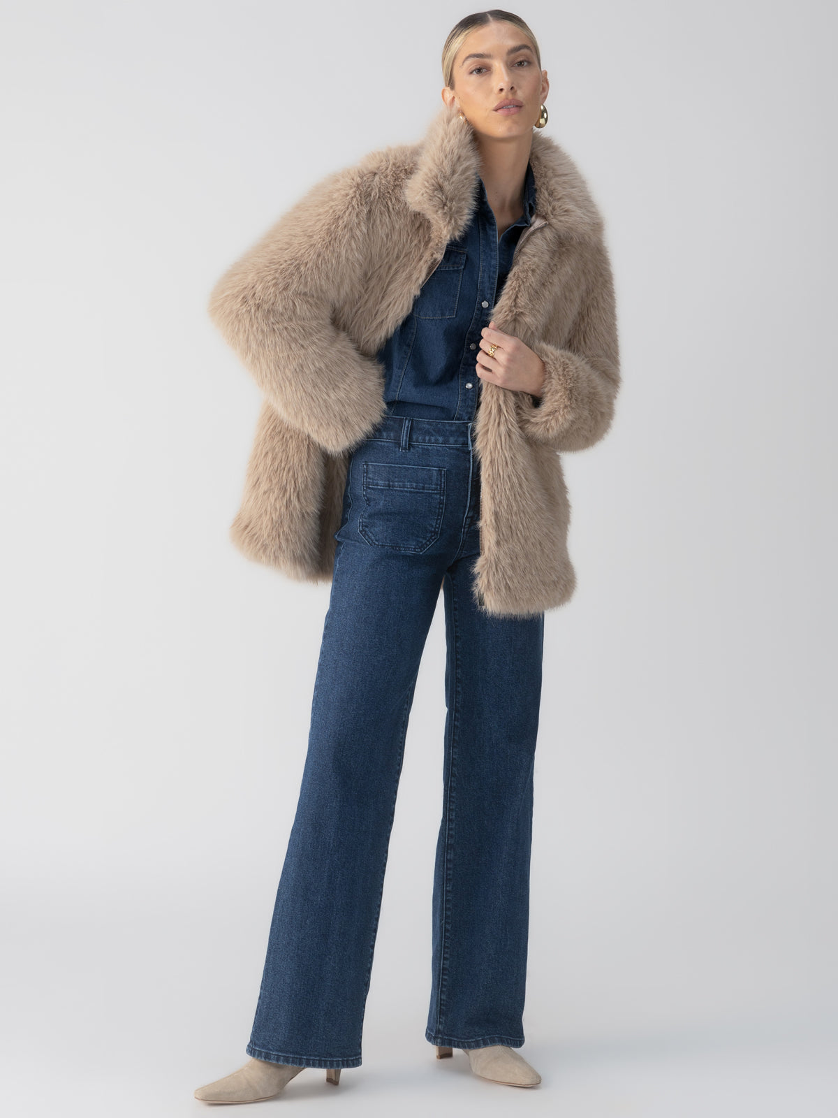 A person stands confidently against a plain background, wearing the Sanctuary Clothing Carmen Fur Coat in Chocolate Sundae over a blue denim jumpsuit. They have their hands in the pockets of the jumpsuit and wear light-colored boots. Their hair is styled back, and they sport hoop earrings.