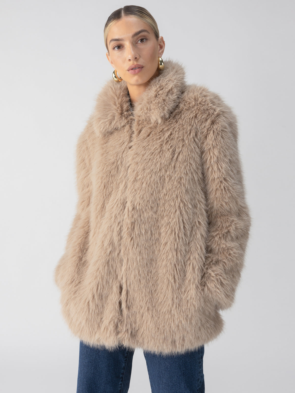 A person wearing the Carmen Fur Coat in Chocolate Sundae by Sanctuary Clothing, which features a high collar and a light beige faux fur texture, stands against a plain background. They have gold hoop earrings and their hands are tucked into the coat pockets. They are looking directly at the camera.