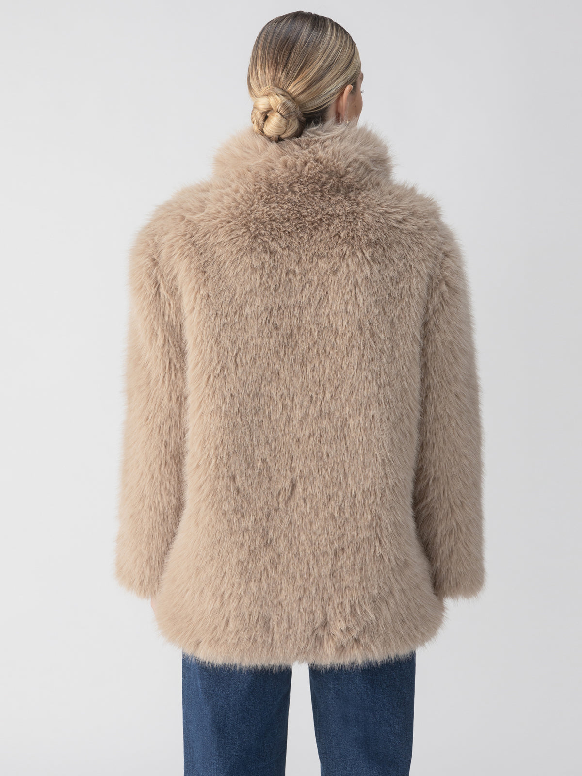 A person with light brown hair in a bun wears the Carmen Fur Coat in Chocolate Sundae by Sanctuary Clothing, appearing from the back. They are dressed in blue jeans. The background is plain and white, providing a clear view of the outfit.