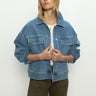 A person with long hair wears Sanctuary Clothing's make way denim jacket wayfarer over a white shirt and olive green pants. They stand against a plain light background, looking to the side with hands gently clasped at the front.