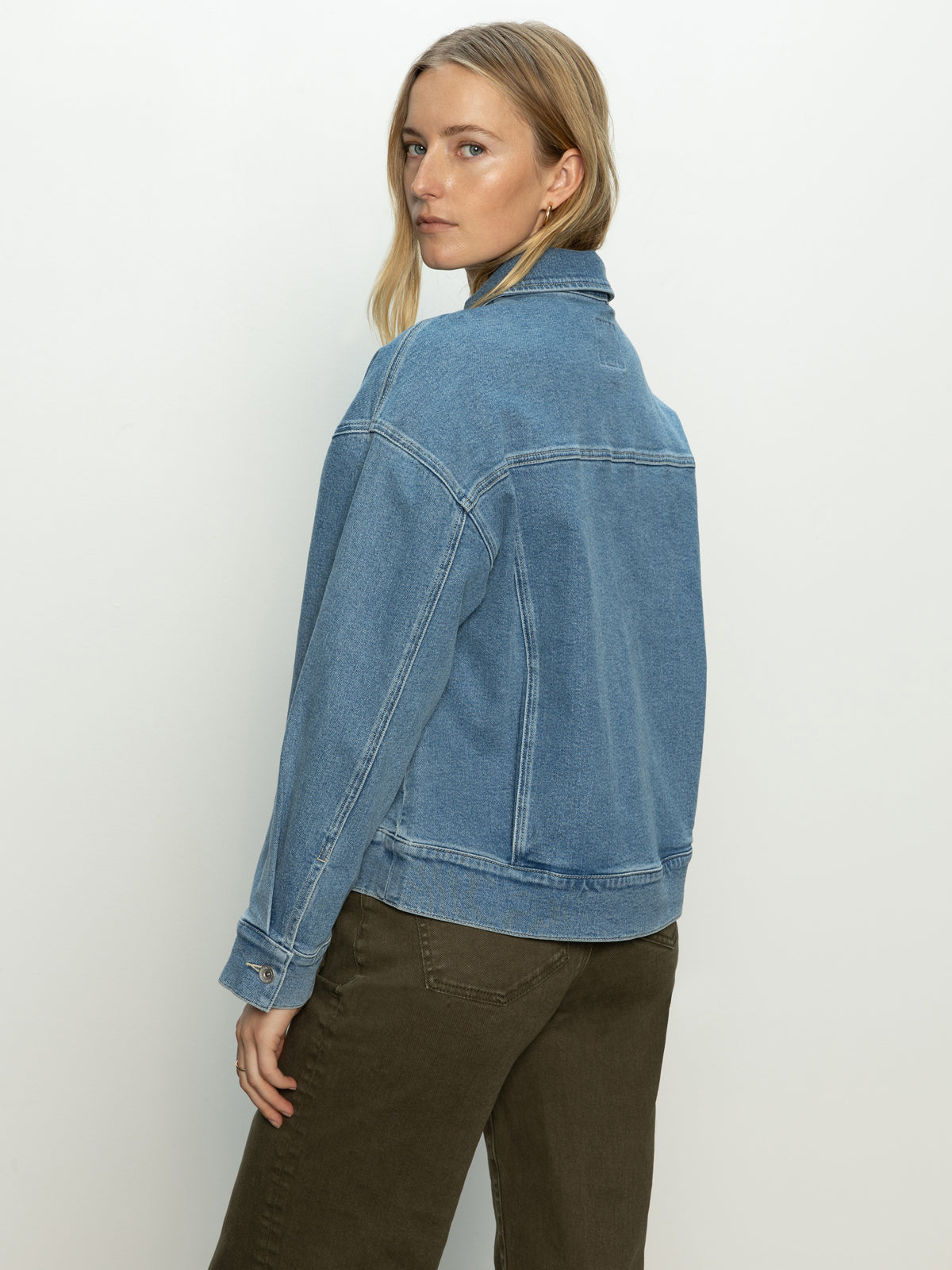 A person with long hair stands against a neutral background, wearing the Sanctuary Clothing make way denim jacket wayfarer and olive green pants, looking over their shoulder.