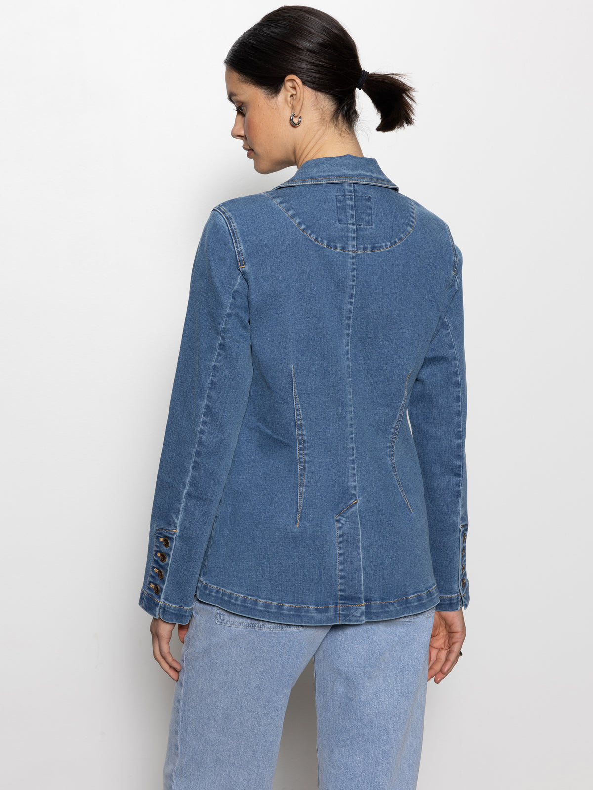 A person with dark hair tied back wears Sanctuary Clothing's sculpted denim blazer best in show, featuring button details on the sleeves. They gaze to the side against a plain white background, paired with blue jeans for a casual look.