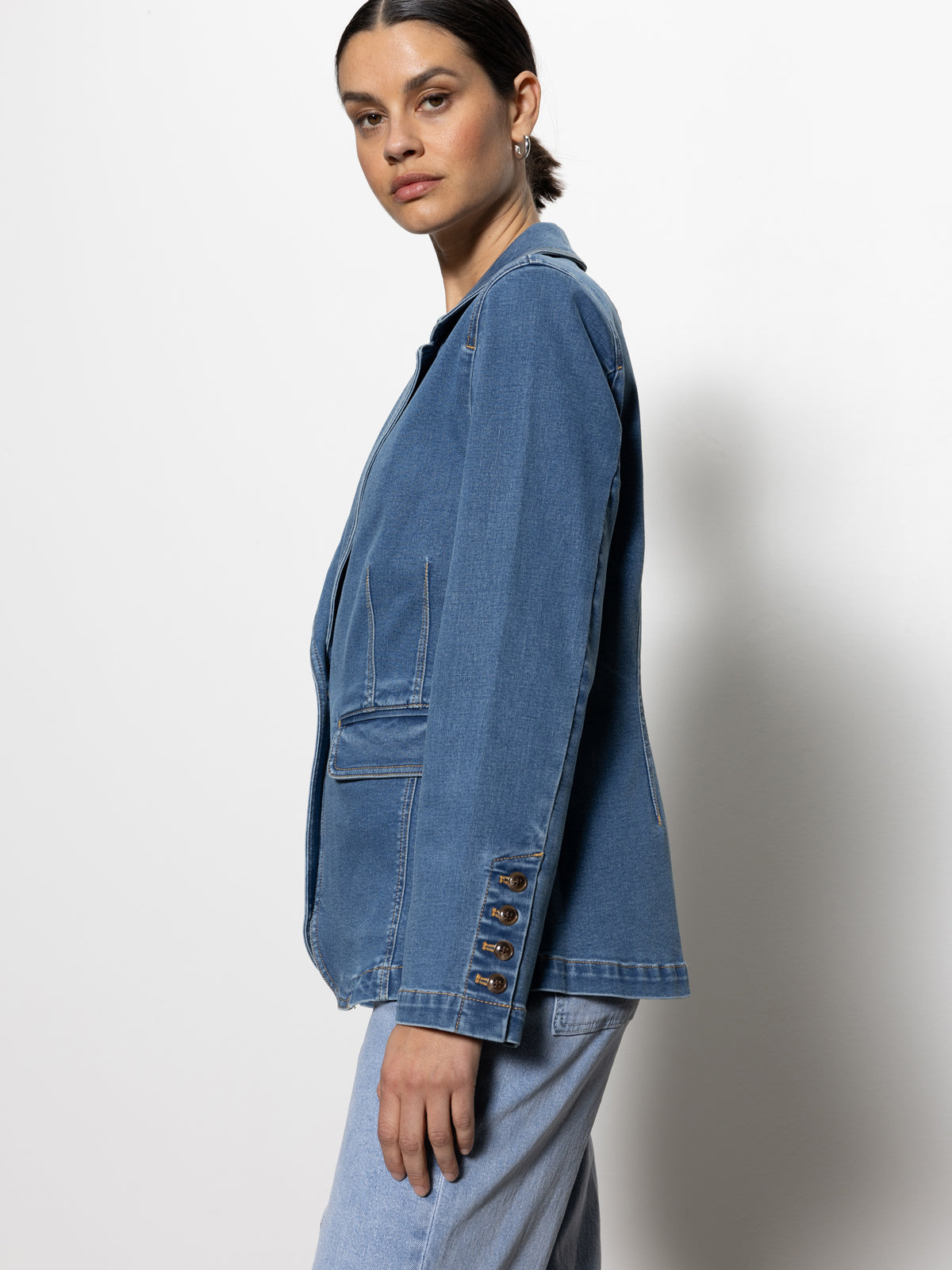 Person wearing the Sculpted Denim Blazer Best in Show by Sanctuary Clothing, featuring buttons on the sleeves and front pockets. They stand against a plain white background, paired with matching denim jeans, looking directly at the camera.