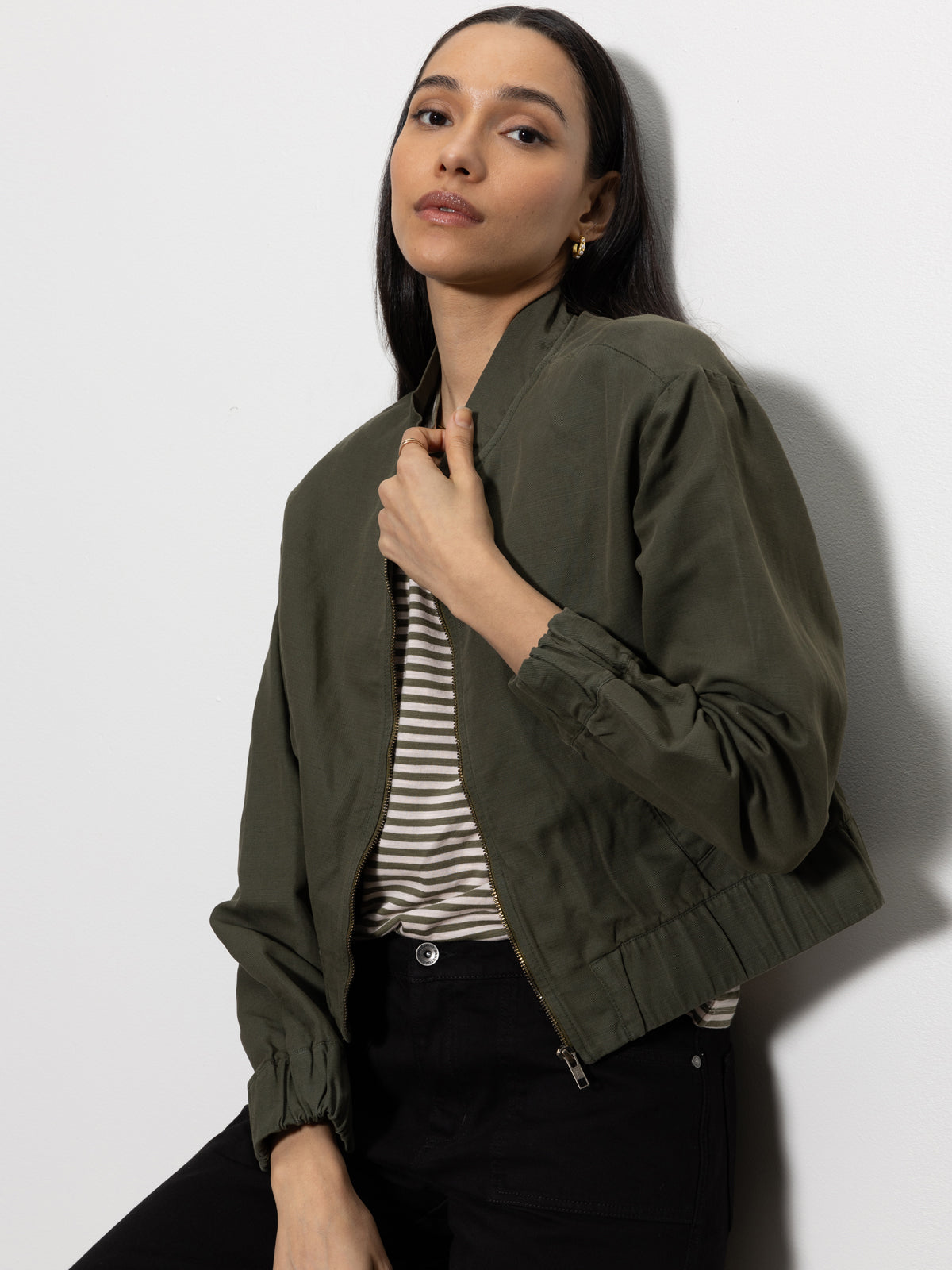 A woman with long dark hair poses against a white wall, wearing Sanctuary Clothing's linen bomber kalamata over a striped shirt and black pants. She lightly holds the jacket's collar with one hand, gazing calmly at the camera.