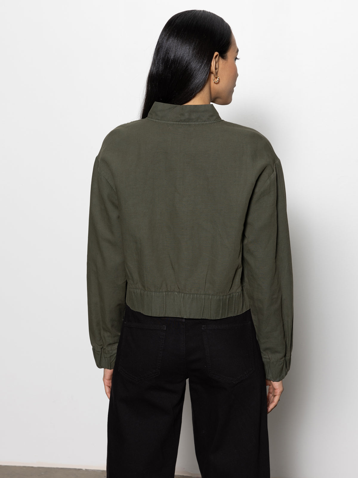 A person with long black hair stands facing away from the camera, wearing a Sanctuary Clothing linen bomber in kalamata green and black pants, against a plain white wall.