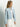 A woman in the Sanctuary Clothing soft moto jacket finish line, a light blue denim piece with zippered pockets and a small collar, stands against a plain background. She pairs it stylishly with a white top and pants, her long straight hair cascading as she gazes to the side.