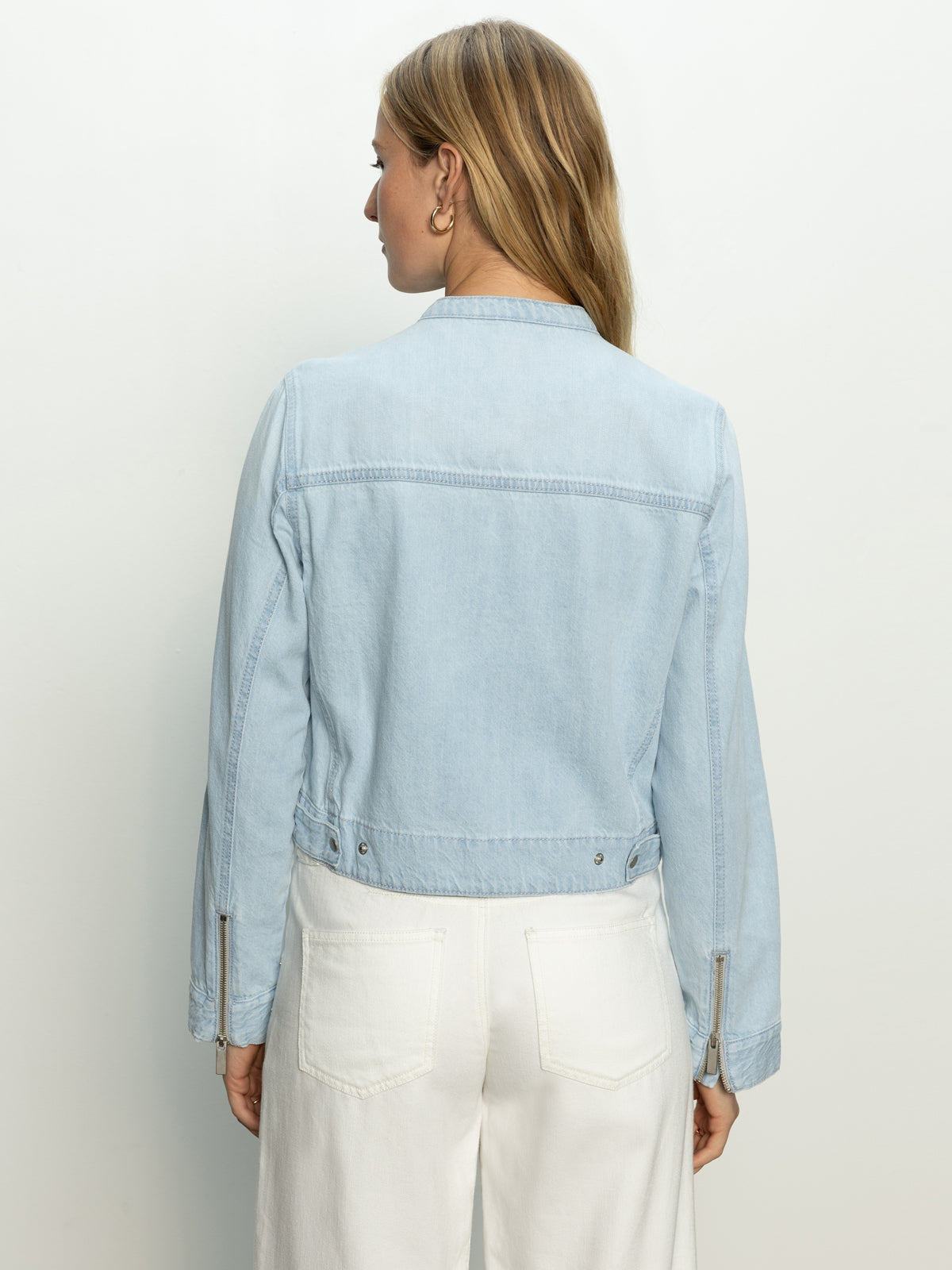 A woman is facing away in a soft moto jacket by Sanctuary Clothing, pairing it with white pants. The light blue denim jacket features button details and zippers on the sleeves, set against a plain, light-colored background.