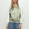 A woman with long blonde hair stands against a neutral background, wearing the Sanctuary Clothing's souvenir jacket in sage—a green and white satin bomber jacket with front zipper and striped cuffs—paired with blue jeans.