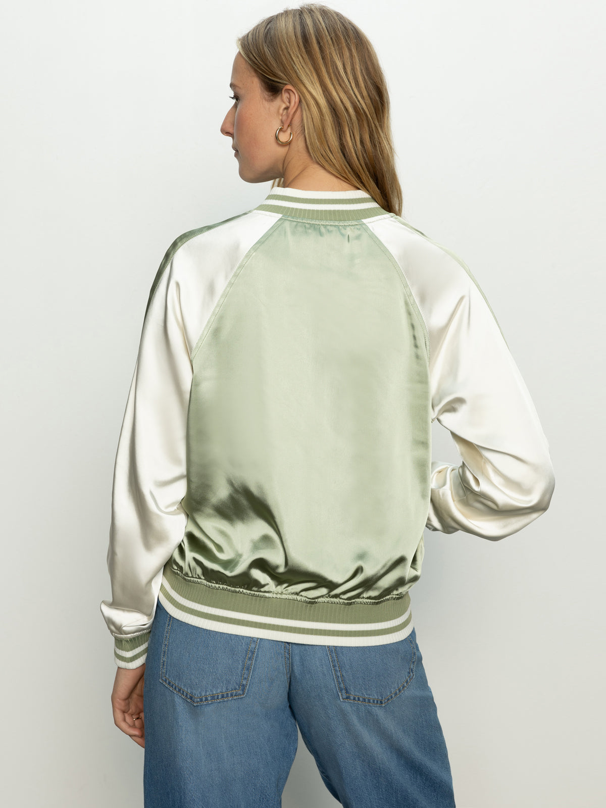 A person with long hair is wearing Sanctuary Clothing's souvenir jacket in sage featuring light green and off-white panels, ribbed collar, cuffs, and waist. They pair it with blue jeans while standing and looking to the side.