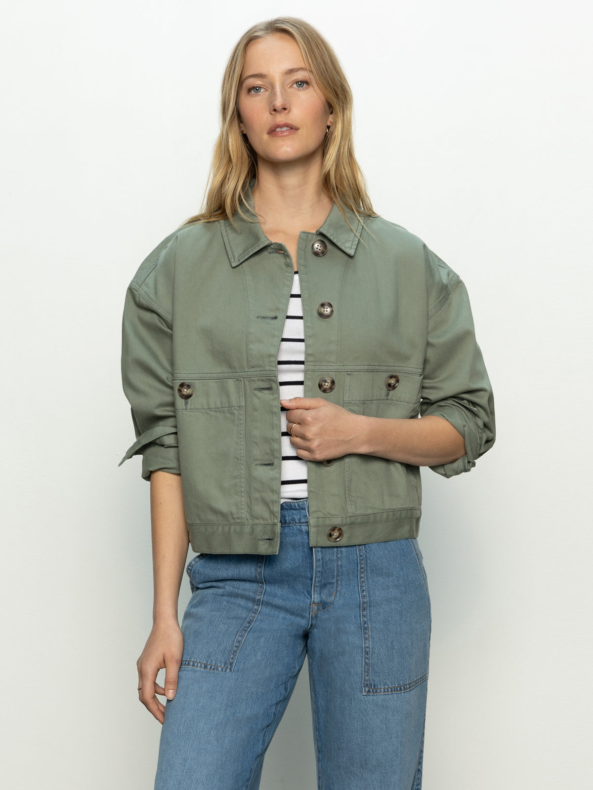 Person in a Sanctuary Clothing coastal denim shacket in dark spruce over a striped shirt and blue jeans, standing against a plain background with relaxed hands, looking forward.