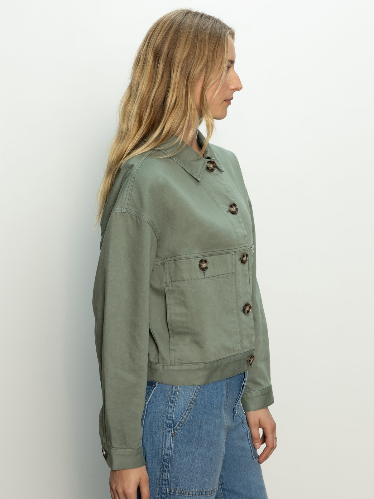 A woman with long blond hair stands in profile wearing the Sanctuary Clothing coastal denim shacket in dark spruce and blue jeans. The shacket features large buttons and front pockets against a plain background.