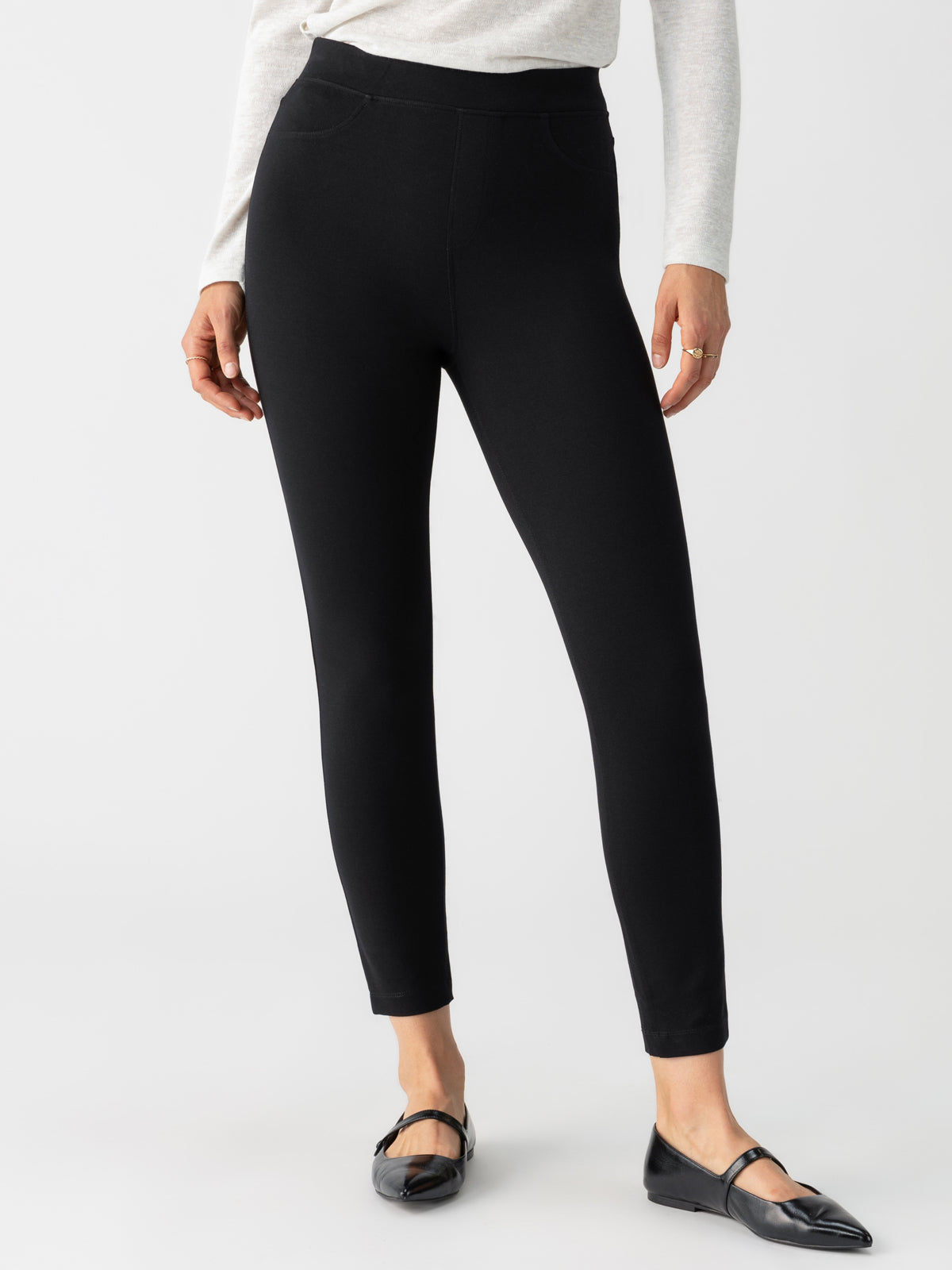 A person wearing the black Runway Legging by Sanctuary Clothing, paired with a light grey, long-sleeved top stands against a plain background. They are also wearing black flats with a slight heel. The photo captures the lower half of the person from the waist to their feet.