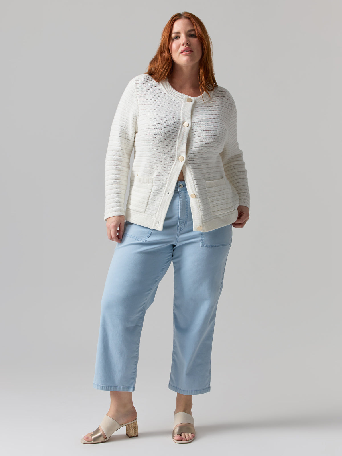 A woman with long red hair is wearing a white button-up cardigan over a light blue top and the Vacation Crop Pant Ultra Pale from the Inclusive Collection by Sanctuary Clothing. She is also wearing beige sandals. The background is plain and light grey.