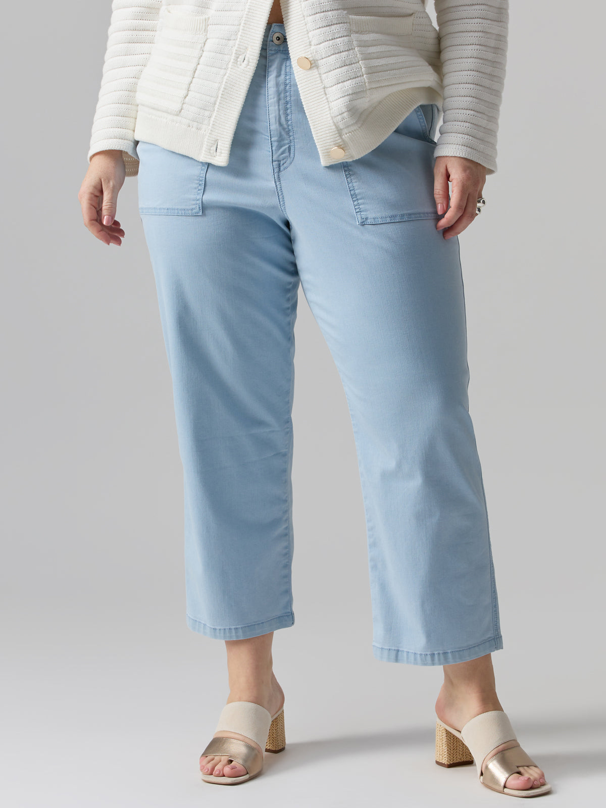 A person wearing Sanctuary Clothing’s Vacation Crop Pant Ultra Pale from the Inclusive Collection, paired with a white textured sweater and beige open-toed block heels, stands against a minimalist grey background. The person's hands are in their pockets, and their head is not visible in the image.