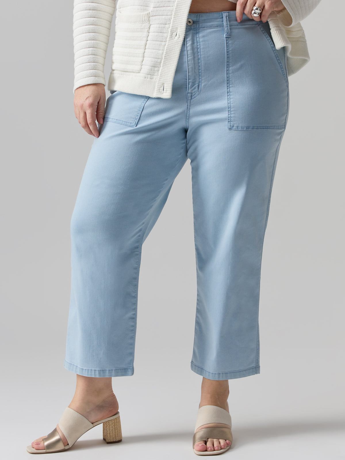 A person wearing the Sanctuary Clothing Vacation Crop Pant Ultra Pale from the Inclusive Collection, a high-waisted, wide-leg design with large front pockets, paired with a white textured cardigan and open-toed beige block-heeled sandals. The photo captures the individual from the chest down to showcase the garments and shoes.