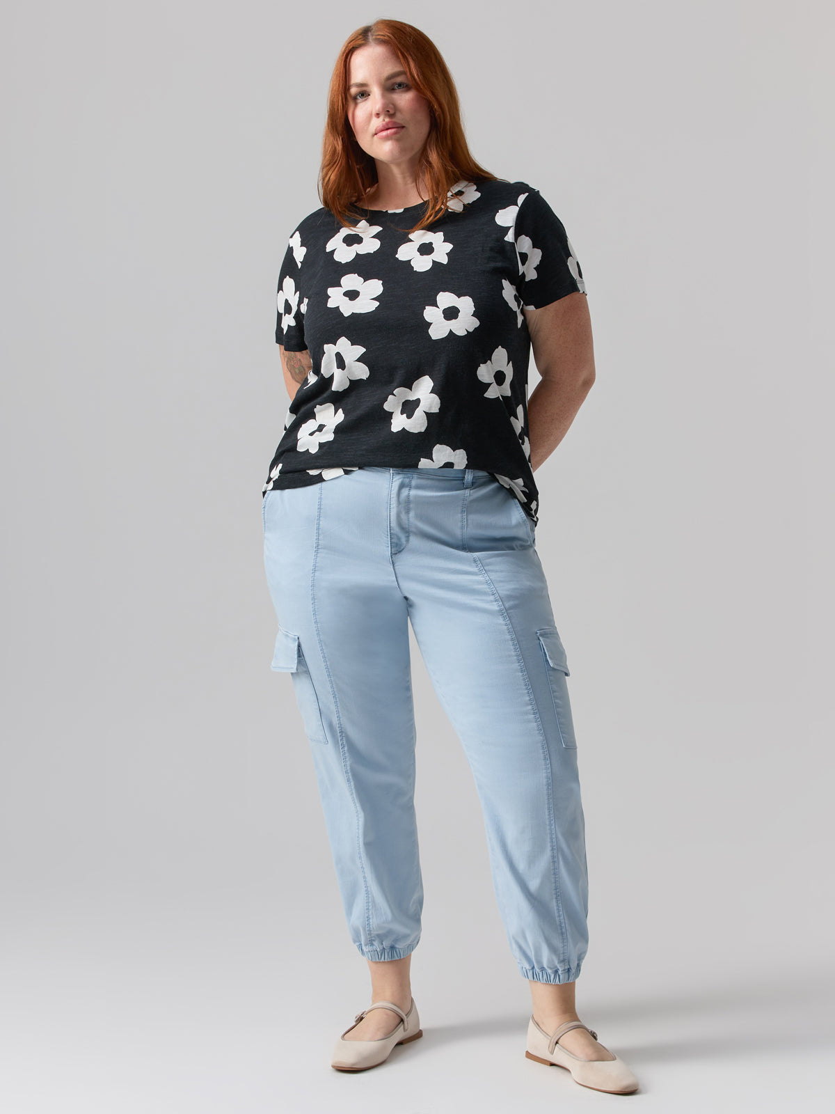 A woman with red hair is wearing a black t-shirt with white flower patterns, Rebel Pant Ultra Pale from Sanctuary Clothing's Inclusive Collection, and beige shoes. She is standing against a plain light gray background, with her arms partially behind her back, looking confidently at the camera.