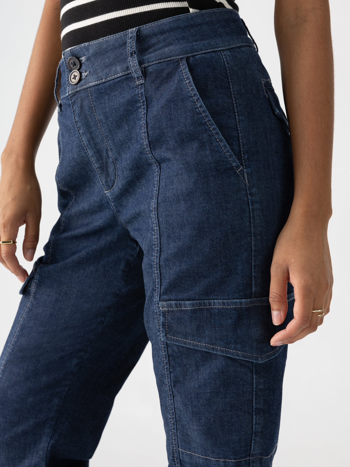 Close-up of a person wearing Sanctuary Clothing's Rebel Pant Bellweather in dark blue denim, paired with a black and white striped top. The pants feature multiple pockets, and the individual is shown from the waist down, with their left hand resting by their side. They are also adorned with rings on their fingers.