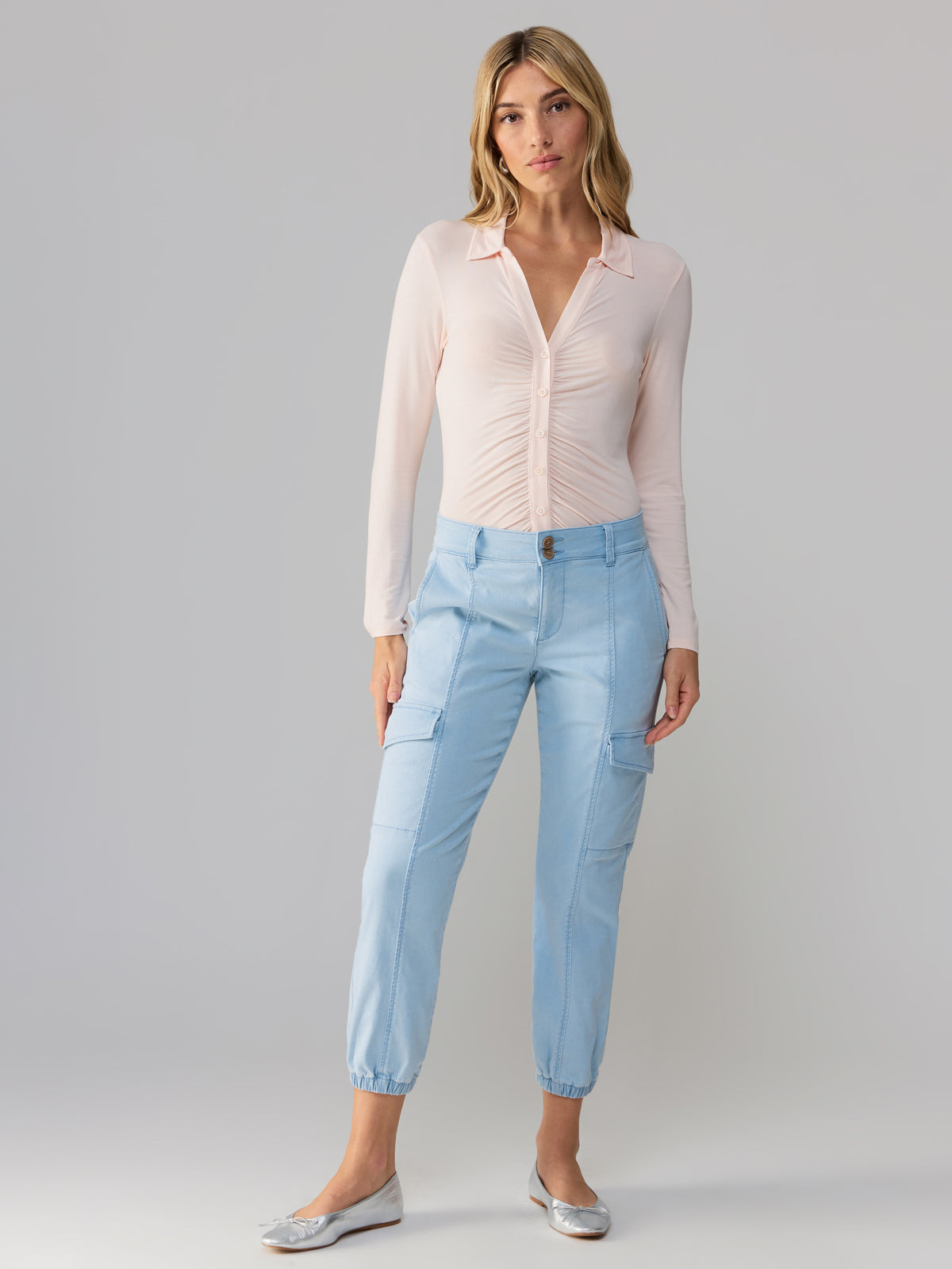 A woman with blonde hair is standing against a neutral background. She is wearing a light pink button-up top with long sleeves and light blue Rebel Pant Ultra Pale cargo-style jogger pants by Sanctuary Clothing. She has a neutral expression and wears silver ballet flats.