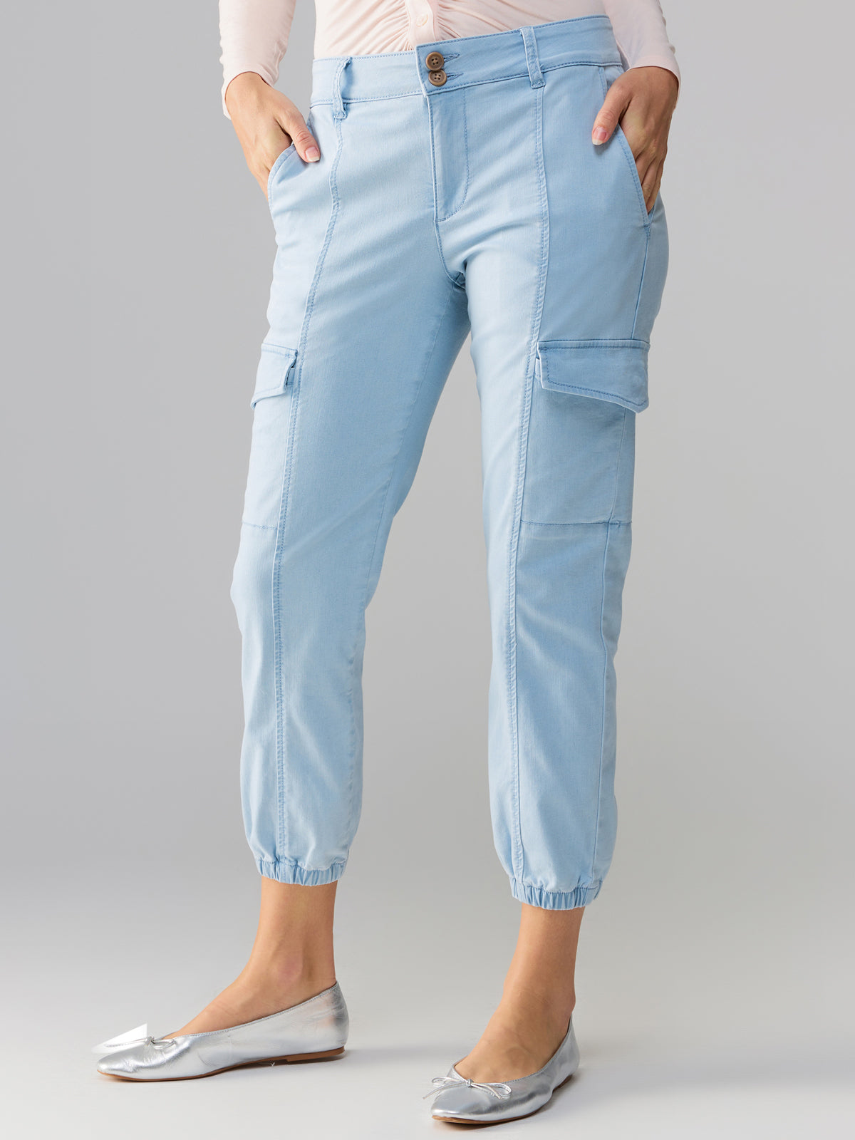 A person wearing the Rebel Pant Ultra Pale from Sanctuary Clothing, light blue cargo pants with side pockets and elastic cuffs, paired with a tucked-in light-colored top and silver flat shoes. The person's hands are in their pockets, standing against a plain background.