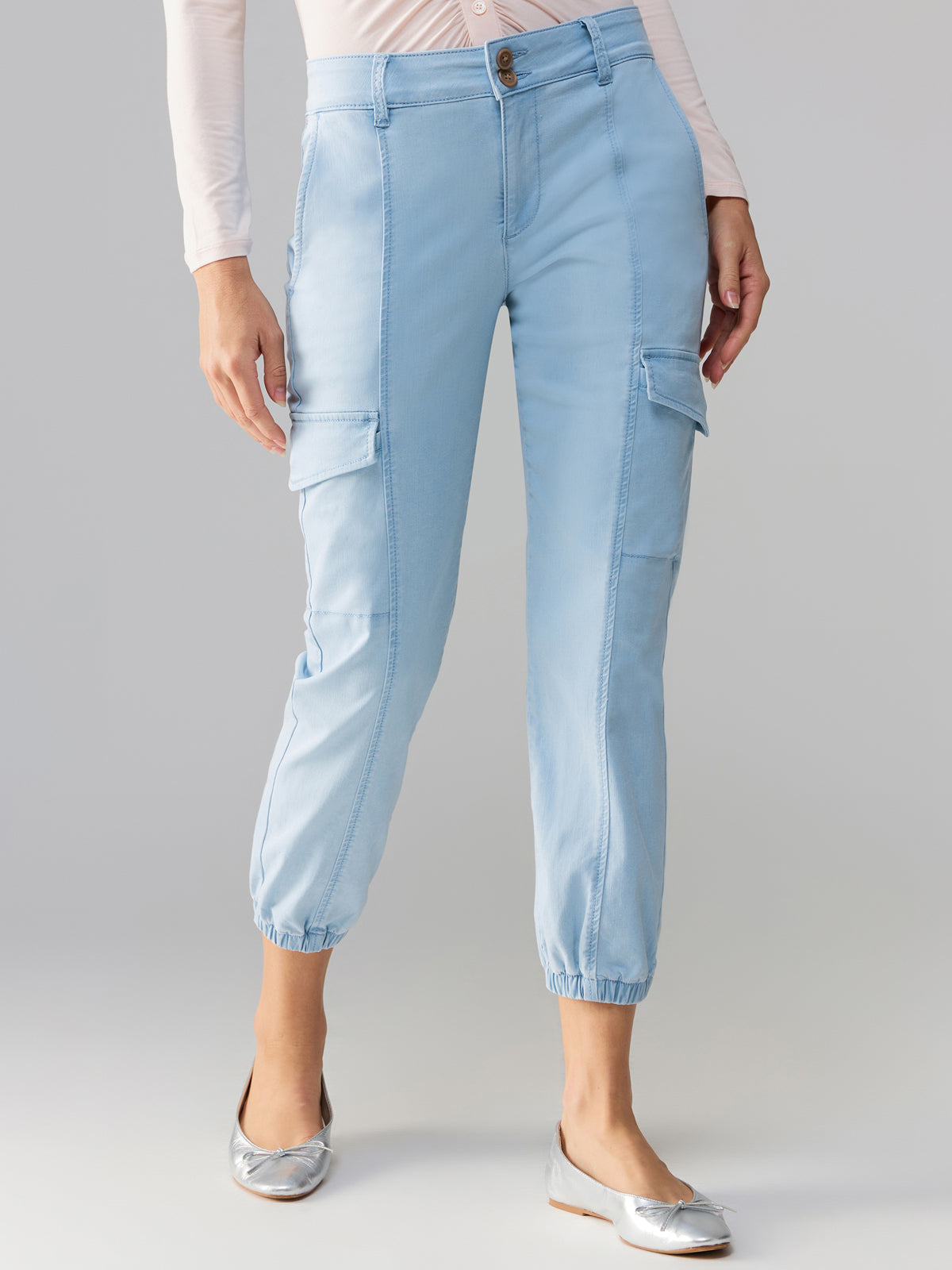 A person wearing the Rebel Pant Ultra Pale from Sanctuary Clothing, featuring side pockets with flaps, two front buttons, and elastic cuffs. They pair it with a light pink long-sleeve top and silver flat shoes.