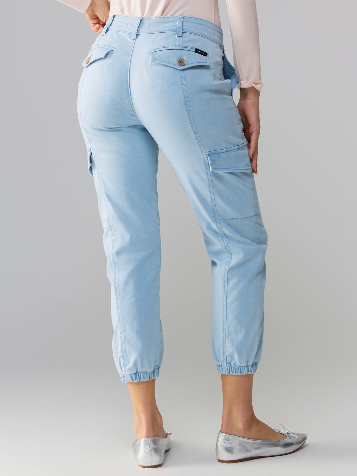 A person is shown from the waist down wearing Sanctuary Clothing's Rebel Pant Ultra Pale, featuring elastic cuffs and multiple pockets. They are also wearing a light-colored long-sleeve top and silver flat shoes against a plain, neutral background.