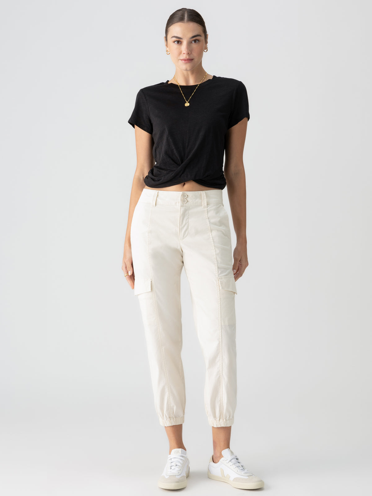 A woman stands facing forward against a plain background. She wears a black, short-sleeved, knotted t-shirt, Sanctuary Clothing's Rebel Pant Eco Natural with elasticated cuffs, white sneakers, and a gold necklace. Her hair is pulled back, and she has a neutral expression.